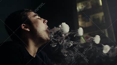 Close-up attractive guy blowing smoke rings in dark hookah room in slow motion
