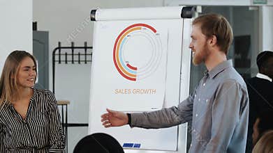 Young professional male business coach leading interactive office seminar, talking to happy female employee at flipchart