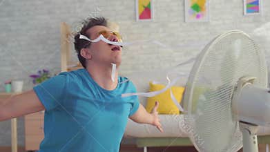 Funny young man with electric fan enjoying cool wind in his apartment slow mo