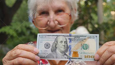 Old woman in eyeglasses showing one hundred dollar bill into camera and smiling outdoor. Happy grandmother holding