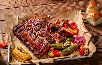 Grilled pork ribs served with grilled corn, salat, bbq sauce, salt pepper and cucumber on parchment paper on a wooden