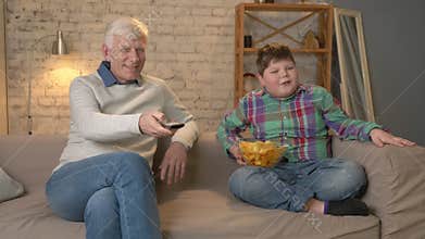 Grandfather and his grandson are sitting on the couch and watching television, eating chips, smiling. An elderly man