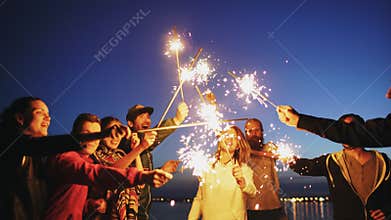 Group of young friends having a beach party. Friends dancing and celebrating with sparklers in twilight sunset