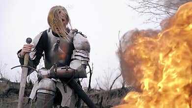 Courageous woman warrior in armor sits leaning on sword against fire