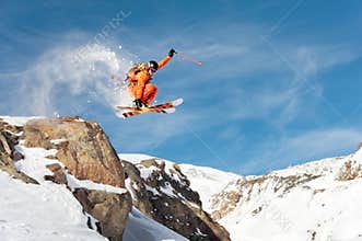 A professional skier makes a jump-drop from a high cliff against a blue sky leaving a trail of snow powder in the