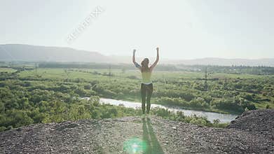 Young sports girl standing on top of the high cliff above the river at beautiful golden sunset, raising her hands up in