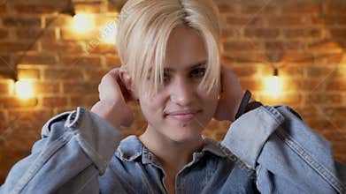 Young cute shot hair girl hipster touches her hair, watching at camera, brick background
