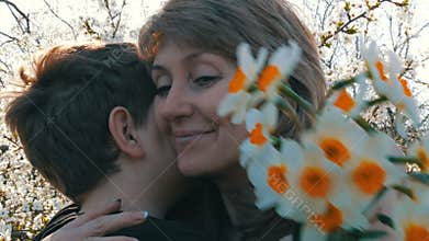 A teenager`s son presents an adult middle-aged mom a bouquet of white daffodils on the background of a blossoming tree