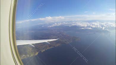 Wing of plane flying high over sea and seashore, sparse white clouds in distance