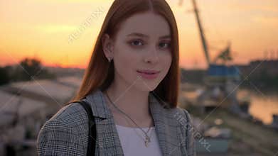Portrait of young beautiful woman with ginger hair standing on bridge and looking into camera, smiling, factory and
