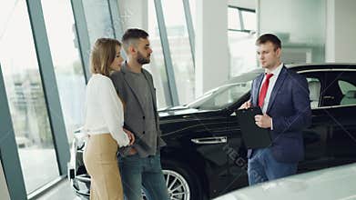 Interested couple man and woman are talking to manager in car dealership while he is telling them about luxury auto