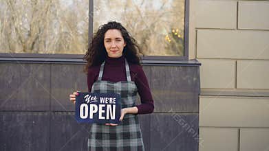 Portrait of attractive confident woman in apron small business owner holding `yes we are open` sign standing outside and
