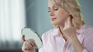 Lady over 50 looking at herself in mirror, rejoicing at result of skin lifting