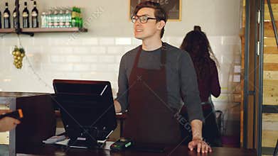 Handsome male cashier is accepting contactless smartphone payments while attractive young waitress is bringing takeaway