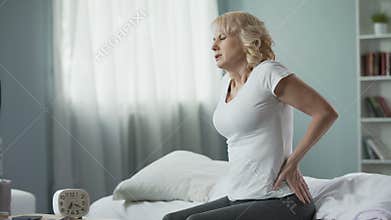 Blond mature lady sitting on bed and touching her back, radiculitis and pain