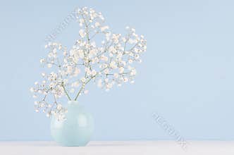 Spring bouquet of white small fluffy flowers in blue smooth circle ceramic vase on soft white wood table and pastel blue wall.