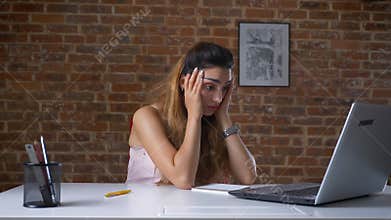 Tired working woman is using her laptop, typing while sitting at desktop over brick wall, bored face