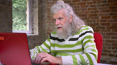 Awesome hairy aged caucasian man with long white beard is typing and demonstrating winning gesture with his fist while