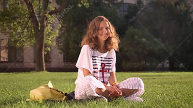 Young smiling caucasian brunette sitting in park on grass, looking in camera, laughing, university in the background