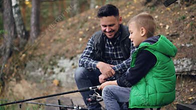 Bearded young man is fishing with cute child on autumn day, boy is holding rod and talking to father learning to catch