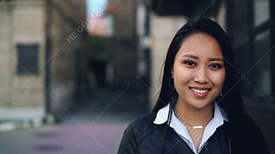 Close-up slow motion portrait of attractive Asian girl looking at camera with happy smile standing in the street wearing