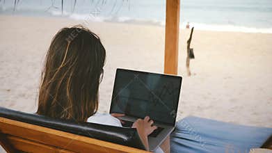 Back view successful CEO businesswoman focused on laptop financial diagrams in lounge chair on exotic ocean resort beach