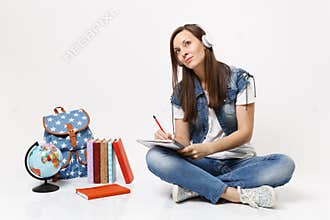 Young pretty woman student in headphones looking up dreaming listen music writing notes on notebook near globe backpack