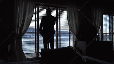 Silhouette of a man talking on cell phone while standing in a hotel room by the window with blinds