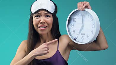 Emotional Asian lady in pajamas and eyemask showing clock, late for work closeup