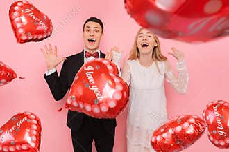 A couple in love, a man and a woman, holding balloons in the shape of a heart on a pink background, enjoy Valentine`s day