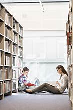 Friends reading book while sitting amidst bookshelves at library