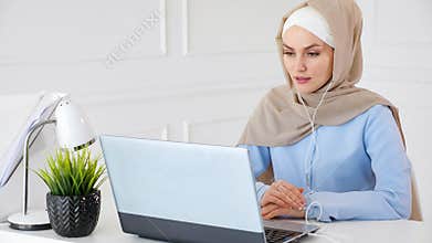 Muslim woman is learning english in earphones online using computer.