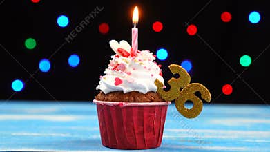 Delicious birthday cupcake with burning candle and number 36 on multicolored blurred lights background