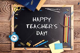 A chalkboard with the text happy teachers day written in it, school supplies and alarm clock on a rustic wooden table