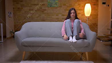 Closeup shoot of young pretty brunette caucasian female being sad depressed and bored sitting on the couch in a cozy