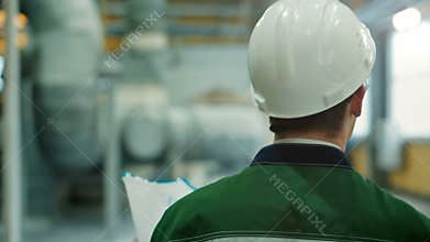 Backview of engineer in hardhat walking in factory and writing down on clipboard