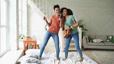 Mixed race young funny girls dance singing with hairdryer and playing acoustic guitar on a bed. Sisters having fun