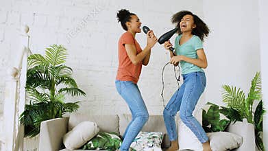 Mixed race young funny girls dance singing with hairdryer and comb jumping on sofa. Sisters having fun leisure in living