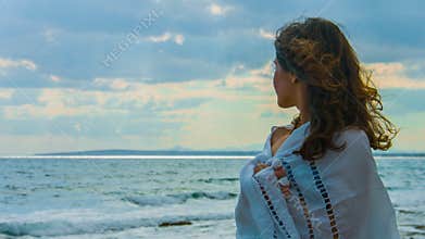 Lonely beautiful woman covered with scarf standing near sea, looking at storm