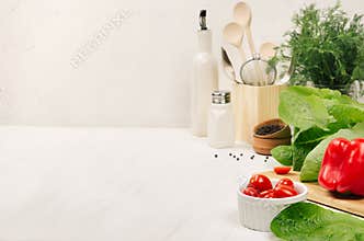 Kitchen white interior with raw fresh green salad, red cherry tomatoes, kitchenware on soft white wood table, copy space.