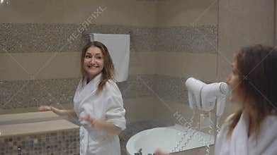 Funny charming girl in white robe took bath and sings in front of mirror with hairdryer.