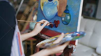 Close-up of artist woman`s hand with brush painting still life picture on canvas in art studio