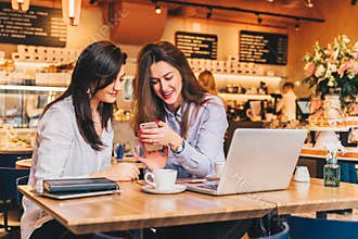 Two young happy women are sitting in cafe at table in front of laptop, using smartphone and laughing.
