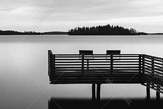 Black and white tranquil scenery of a lake with pier and two chairs