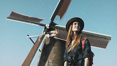 Portrait of traveller woman near old windmill. Incredible cinematic low angle. Girl in hat in front of farm mill. 4K