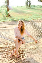 Young pretty blonde female person sitting on wicker hammock, sand background.