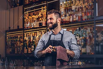 Cheerful stylish brutal barman is cleaning the glass with a cloth at bar counter background.