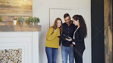 Cheerful realtor is meeting beautiful young couple in new house, opening door, showing documents and talking to clients