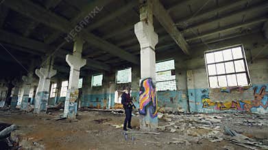 Male graffiti artist in respirator is shaking spray paint then painting on high pillar inside dirty empty building