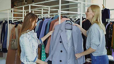 Young woman is helping her friend to find coat. She is bringing her new model, telling about it and showing its